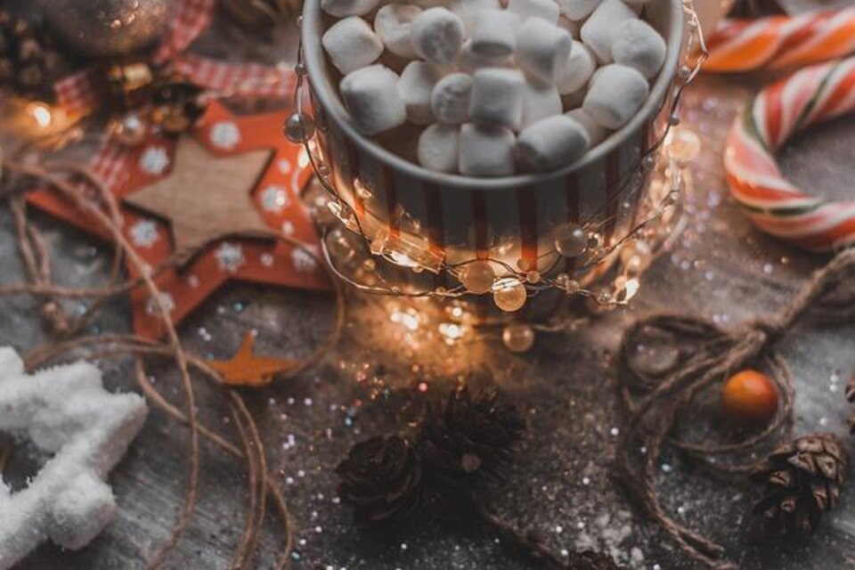 Eine Tasse mit Schokolade und vielen Marshmallows steht auf einem festlich dekorierten Tisch. Drumherum liegen Weihnachtssterne, Tannenzapfen und Lichterketten. | © pixabay