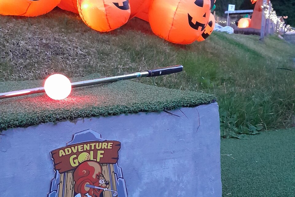 Eine bunte Minigolfanlage mit leuchtenden Kürbissen und einem Schild, das die Nummer eins anzeigt. Die Umgebung ist grün und von Bäumen umgeben. | © Mountain Adventure Golf