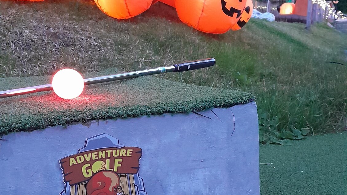 Eine bunte Minigolfanlage mit leuchtenden Kürbissen und einem Schild, das die Nummer eins anzeigt. Die Umgebung ist grün und von Bäumen umgeben. | © Mountain Adventure Golf