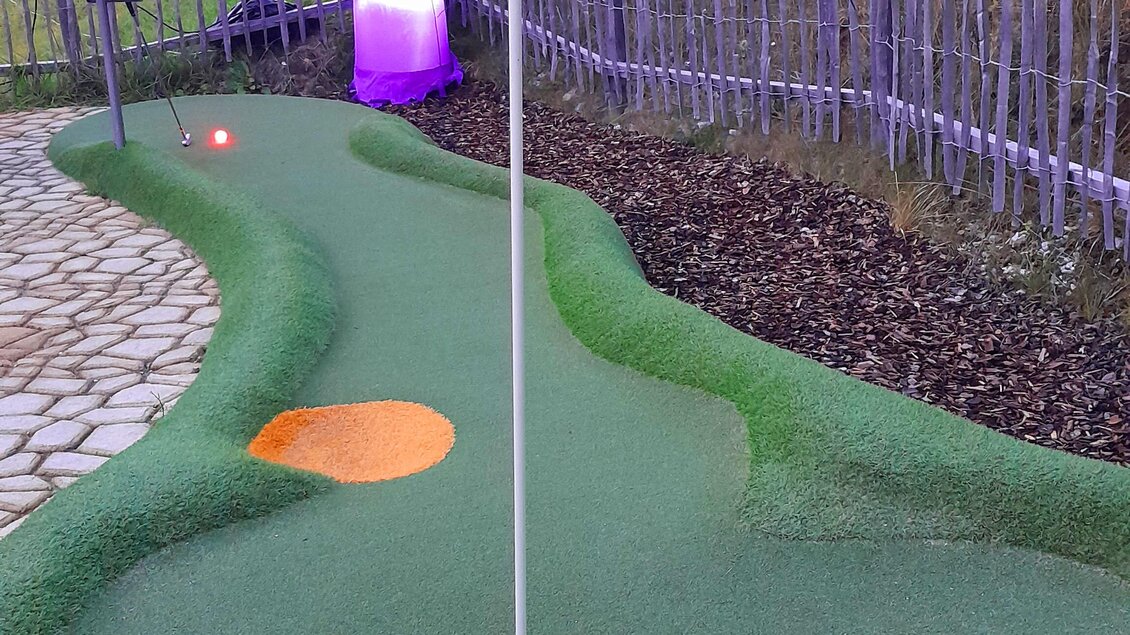Eine Minigolfbahn mit einem Loch und einer Fahne. Im Hintergrund steht eine leuchtende Figur. | © Mountain Adventure Golf