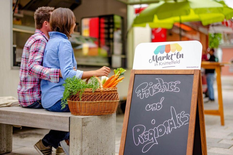 Eine gemütliche Marktszene mit einem Paar, das auf einer Bank sitzt. Vor ihnen steht ein Korb mit frischem Gemüse und ein Schild mit der Aufschrift „Frisches und Regionales“. | © Stadtgemeinde Knittelfeld
