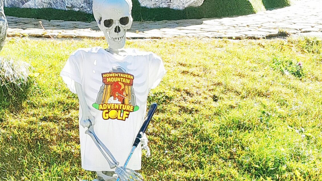 Ein Skelett sitzt auf einer Bank und trägt ein T-Shirt. Im Hintergrund sind Grüns und einige Hindernisse zu sehen. | © Mountain Adventure Golf