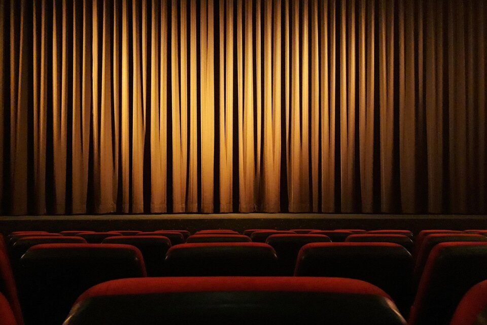 Ein leerer Theatersaal mit roten Sitzen und einem dunklen Vorhang. Das Licht beleuchtet den Vorhang in sanften goldenen Tönen. | © Pixabay